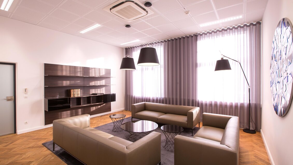 Ein modernes Büro mit hellen Wänden und Holzfußboden. Die Einrichtung umfasst zwei bequeme Sofas, einen Glastisch und große Fenster mit Vorhängen.