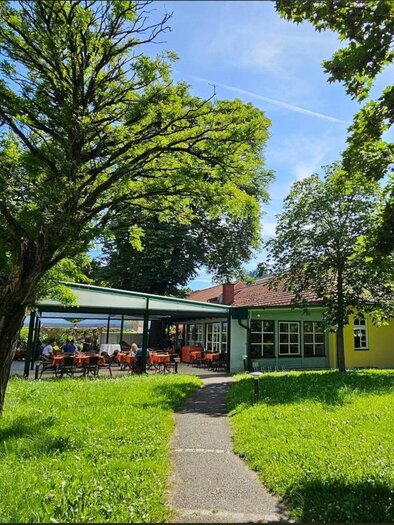 Ein einladendes Café unter Bäumen mit einer Terrasse im Freien. Der Himmel ist klar und die Umgebung ist grün und gepflegt. | © Kastanienlaube