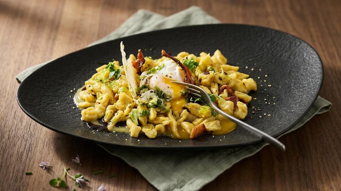 Steirische Carbonara mit Spätzle, Ei, Speck und Kräutern auf einem schwarzen Teller. | © Gabriel Ortet Boll