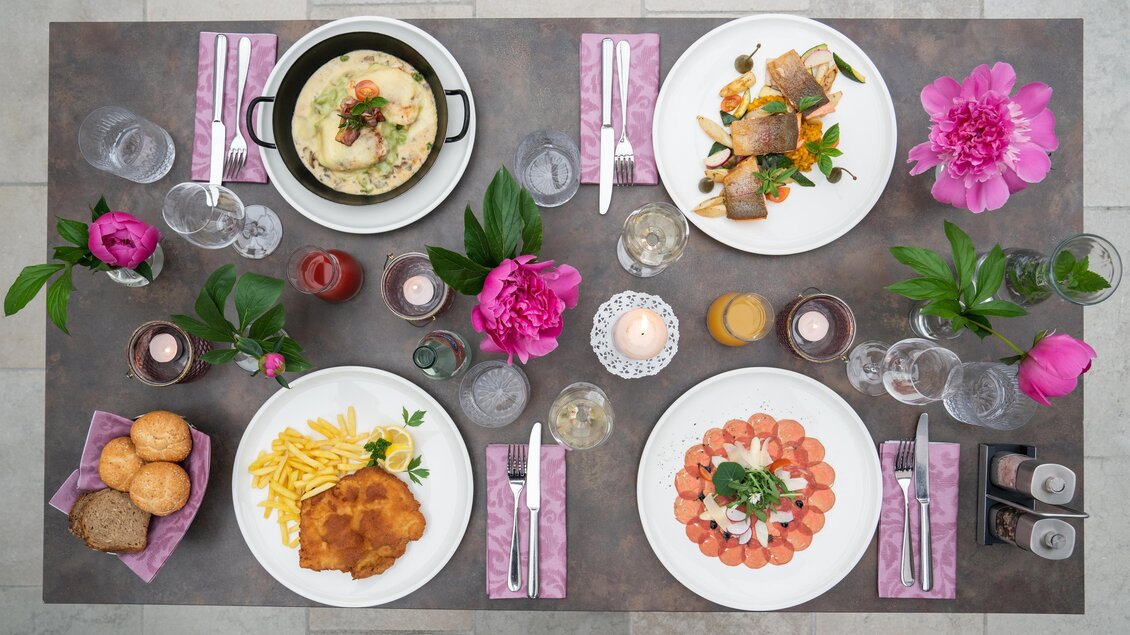 Ein schön gedeckter Tisch mit mehreren Gerichten, darunter Fisch, Fleisch und Pasta. Frische Blumen und Kerzen verleihen dem Bild eine einladende Atmosphäre. | © Gollner