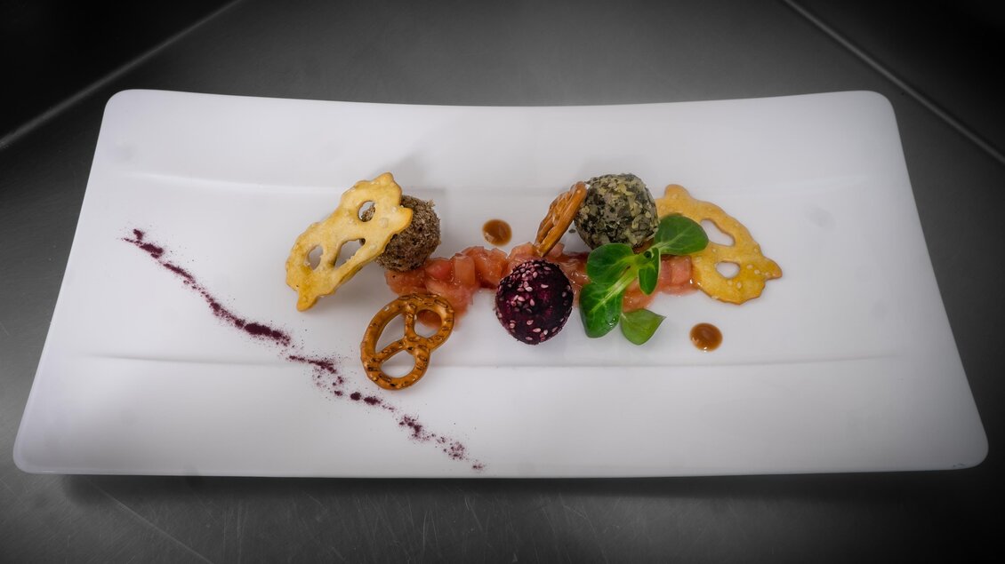 Ein kunstvoll angerichteter Teller mit zwei frittierten Bällchen, einer Garnitur aus getrockneten Gemüsescheiben und einem farblichen Sauce-Tupfer. Frische Kräuter und knusprige Brezeln ergänzen das Gericht. | © Gollner