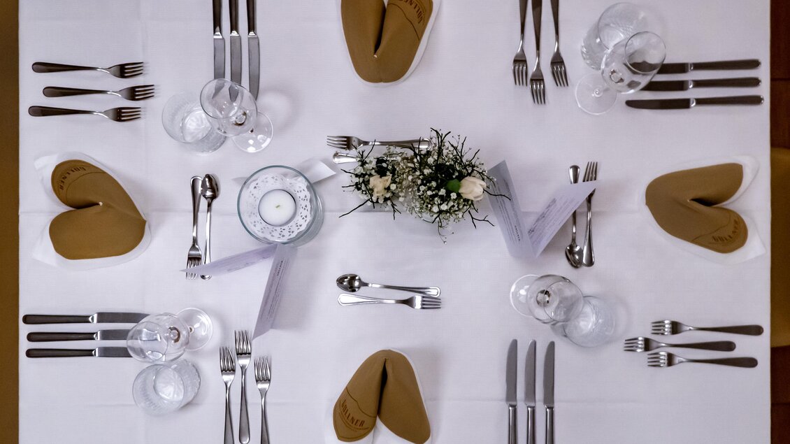 Ein schön gedeckter Tisch mit weißen Tischdecken und eleganten Bestecken. In der Mitte befindet sich ein Blumenarrangement, und die Servietten sind in Herzform gefaltet. | © Gollner