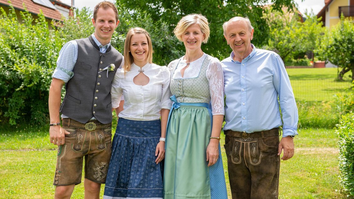 Eine Gruppe von vier Personen in traditioneller bayerischer Tracht steht auf einer grünen Wiese. Im Hintergrund sind Bäume und Gebäude zu sehen. | © Hotel-Restaurant Gruber
