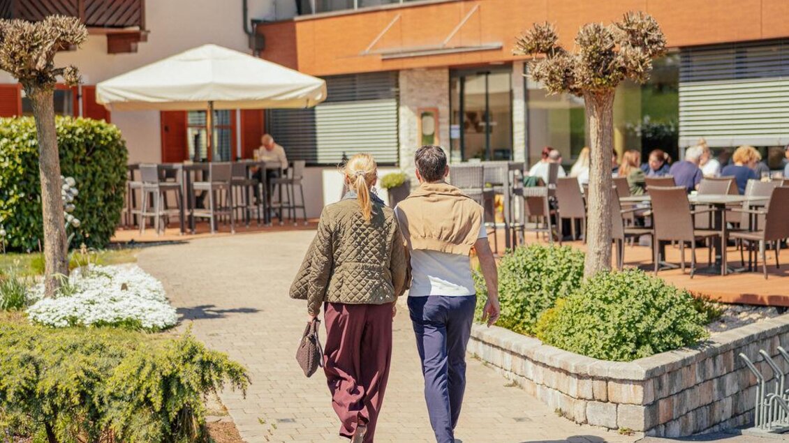 Zwei Personen gehen auf einem gepflasterten Weg zu einem Restaurant im Freien. Im Hintergrund sind Stühle und Tische unter Sonnenschirmen zu sehen. | © N.Ulrich