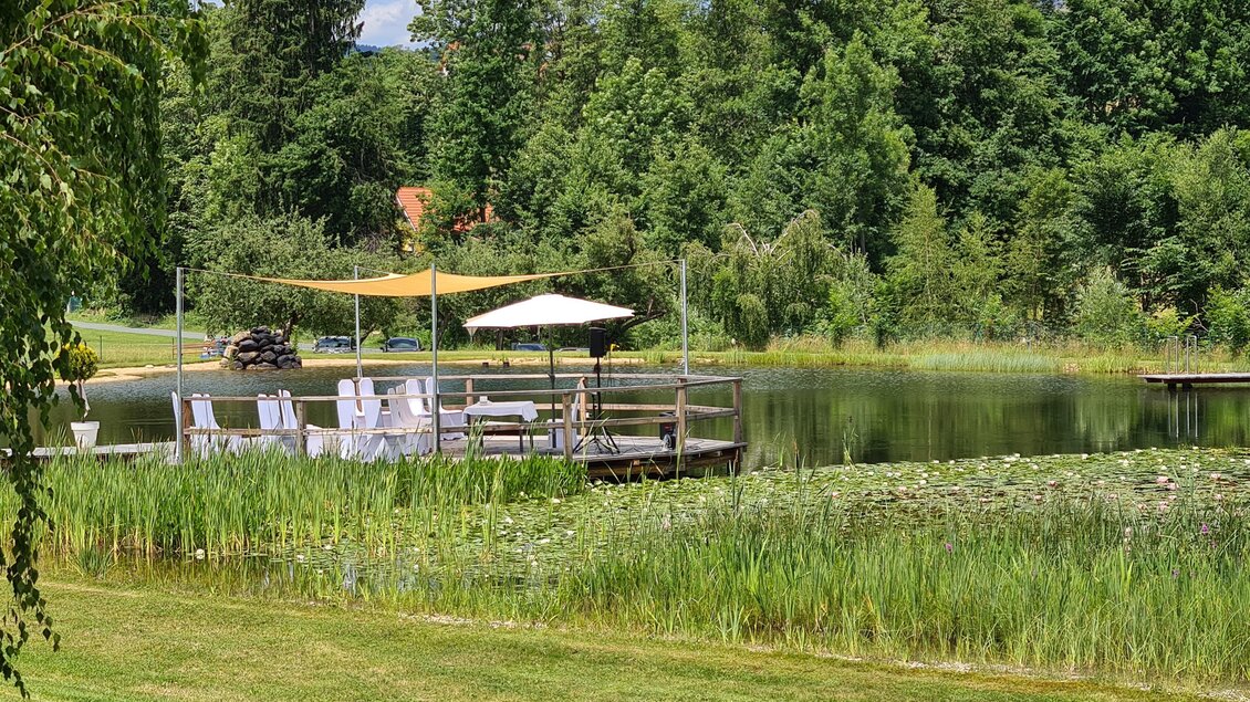 Ein ruhiger See mit einer Holzterrasse und Sonnenliegen. Umgeben von grünen Bäumen und einer schönen Wiese. | © Hotel-Restaurant Gruber