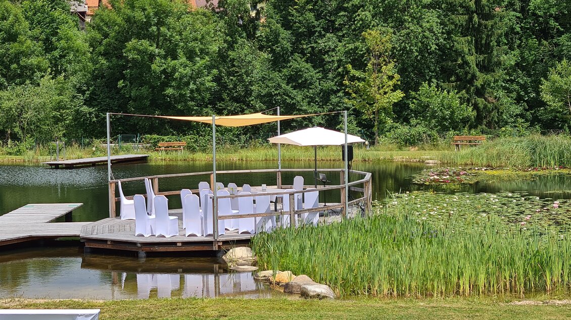 Ein ruhiger Teich mit einer Holzterrasse, auf der Weißes wie in einem feierlichen Arrangement steht. Umgeben von üppigem Grün und Bäumen, strahlt der Ort eine friedliche Atmosphäre aus. | © Tourismusverband Oststeiermark