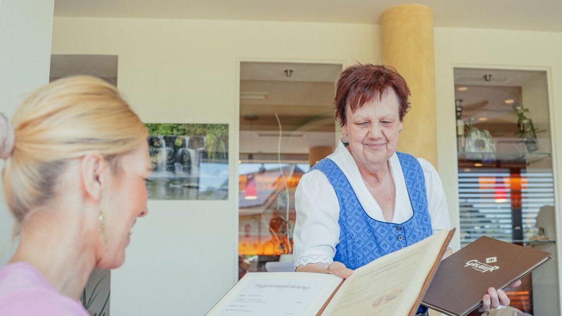 Eine Frau schaut sich das Menü in einem Restaurant an. Eine Bedienung in traditioneller Tracht bringt weitere Informationen. | © N. Ulrich