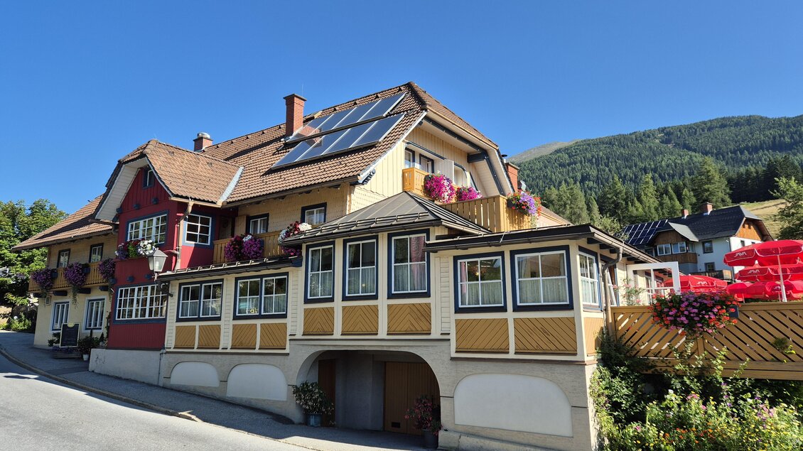 Ein charmantes Haus mit bunten Blumenfenstern und Sonnenkollektoren. Im Hintergrund sind grüne Berge und ein klarer blauer Himmel zu sehen. | © TV Murau