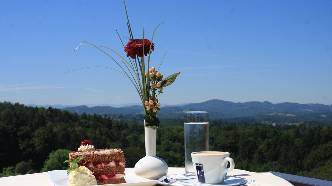 Ein köstliches Stück Torte mit Sahne und ein Kaffee stehen auf einem Tisch. Im Hintergrund erstreckt sich eine malerische Aussicht auf Hügel und Wälder. | © Restaurant Hasenwirt