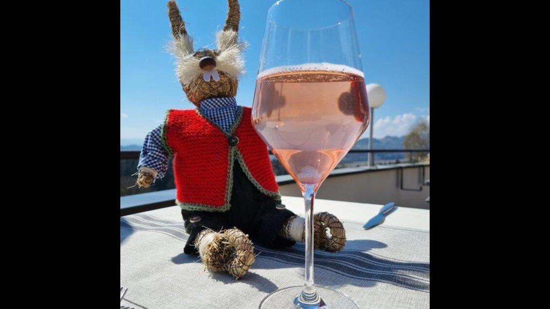 Ein gehäkelter Hase sitzt auf einem Tisch und trägt eine rote Weste. Neben ihm steht ein Glas mit rosa Getränk. | © Restaurant Hasenwirt