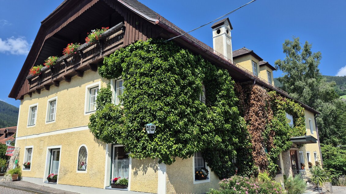 Ein charmantes, gelbes Gebäude mit begrünten Wänden und einem Walmdach. Die Fassade ist mit Pflanzen geschmückt und umgeben von einer malerischen Landschaft. | © TV Hochsteiermark