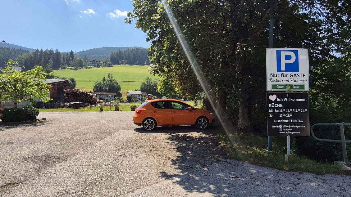 Ein Parkplatz in der Natur mit einem orangefarbenen Auto. Im Hintergrund sieht man Wiesen und Wälder unter einem blauen Himmel. | © TV Hochsteiermark