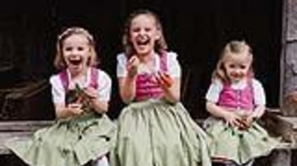Drei fröhliche Mädchen in traditionellen Trachten sitzen auf einer Holzbank. Sie lachen und halten kleine Gegenstände in den Händen.