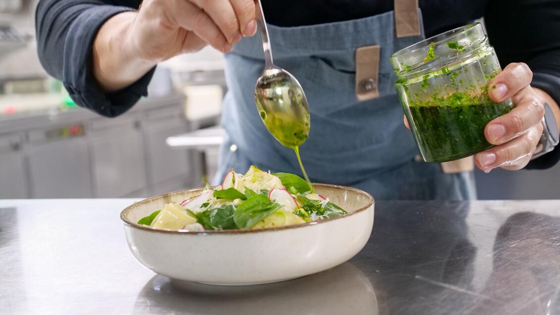 Ein Koch verleiht einem Salat mit frischen Kräutern den letzten Schliff. Das Gericht wird in einer weißen Schüssel präsentiert, während ein Glas mit grüner Soße in der Hand gehalten wird. | © Kirchenwirt Stubenberg