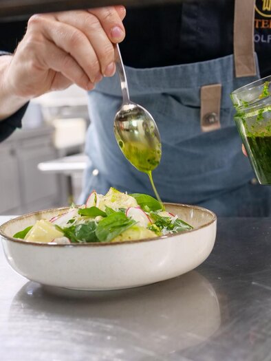 Ein Koch verleiht einem Salat mit frischen Kräutern den letzten Schliff. Das Gericht wird in einer weißen Schüssel präsentiert, während ein Glas mit grüner Soße in der Hand gehalten wird. | © Kirchenwirt Stubenberg