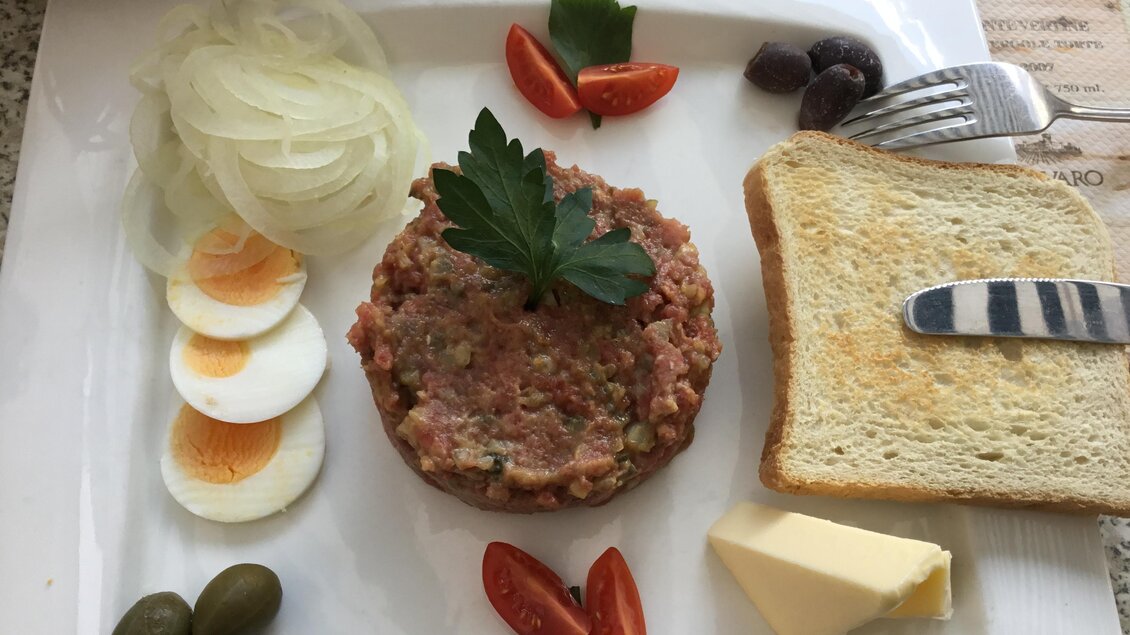 Ein Teller mit Tatar, garniert mit frischer Petersilie, Zwiebeln und Kirschtomaten. Dazu gibt es hartgekochte Eier, Oliven, Kapern und geröstetes Brot mit Butter. | © Landhotel  Groggerhof