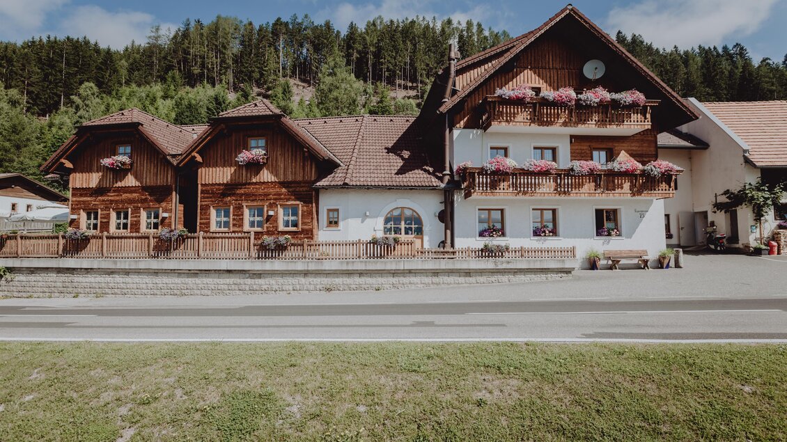 Ein traditionelles Holzhaus mit Balkonen und Blumenarrangements. Im Hintergrund sind Bäume und ein blauer Himmel zu sehen. | © Karoline Karner