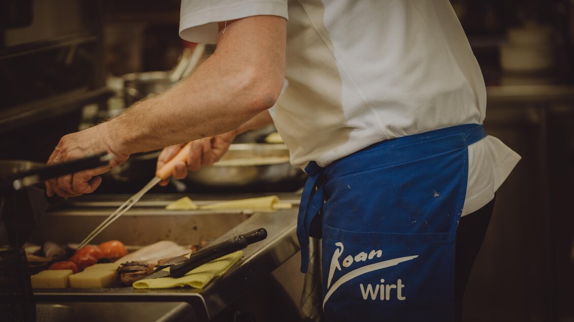 Ein Koch bereitet Lebensmittel in einer Restaurantküche vor. Er trägt eine Schürze mit dem Schriftzug „Wirt“. | © Karoline Karner