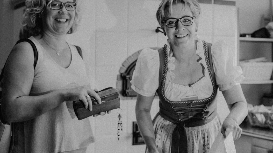 Zwei Frauen lächeln und stehen in einer hellen Küche. Eine trägt ein traditionelles Dirndl und die andere hält eine Geldbörse in der Hand. | © Karoline Karner