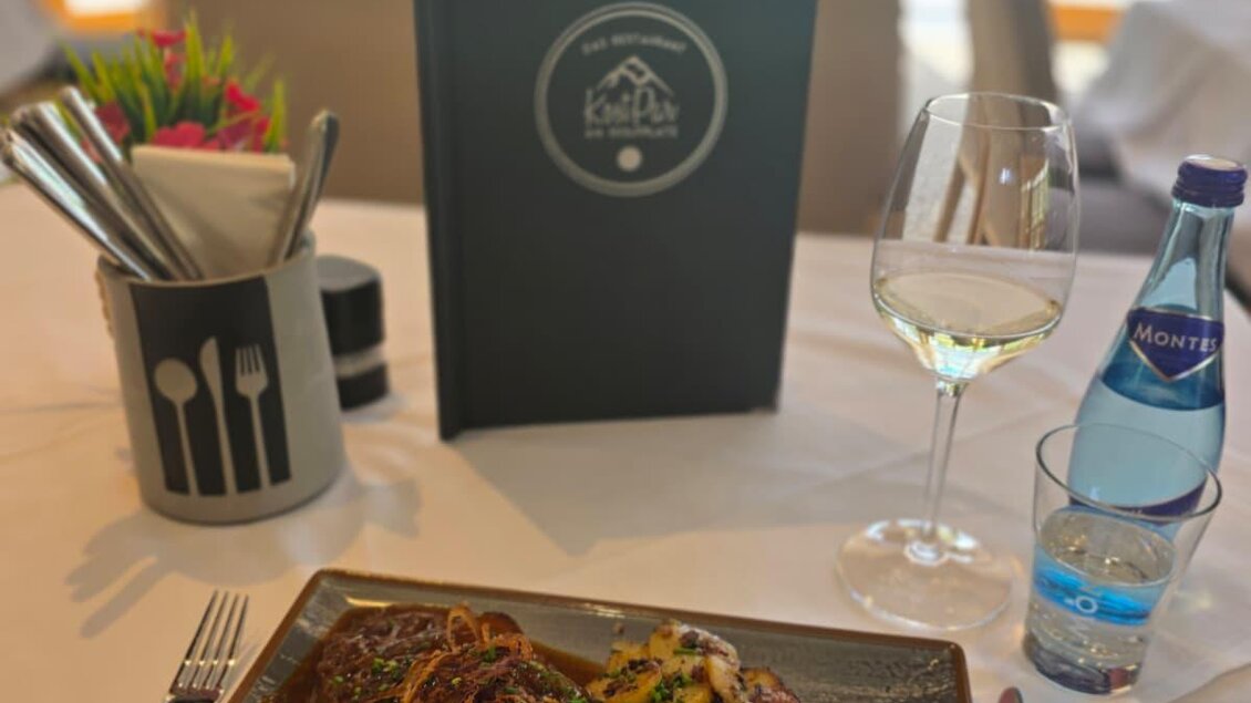 Ein köstliches Gericht mit Fleisch, Kartoffeln und grünem Gemüse auf einem eleganten Teller. Im Hintergrund sind eine Wasserflasche und ein Menü sichtbar.