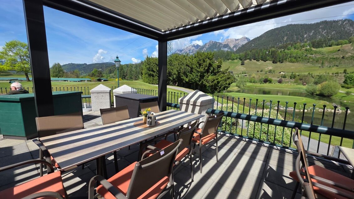 Eine gemütliche Terrasse mit einem großen Tisch und Stühlen. Im Hintergrund sind grüne Wiesen und Berge zu sehen.