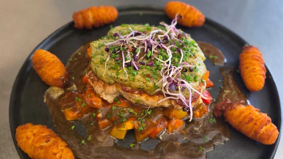 Ein Teller mit einer bunten Mahlzeit, die aus Gemüse, einer grünen Sauce und knusprigen Beilagen besteht. Um das Hauptgericht sind orangefarbene Kartoffelknödel angeordnet.