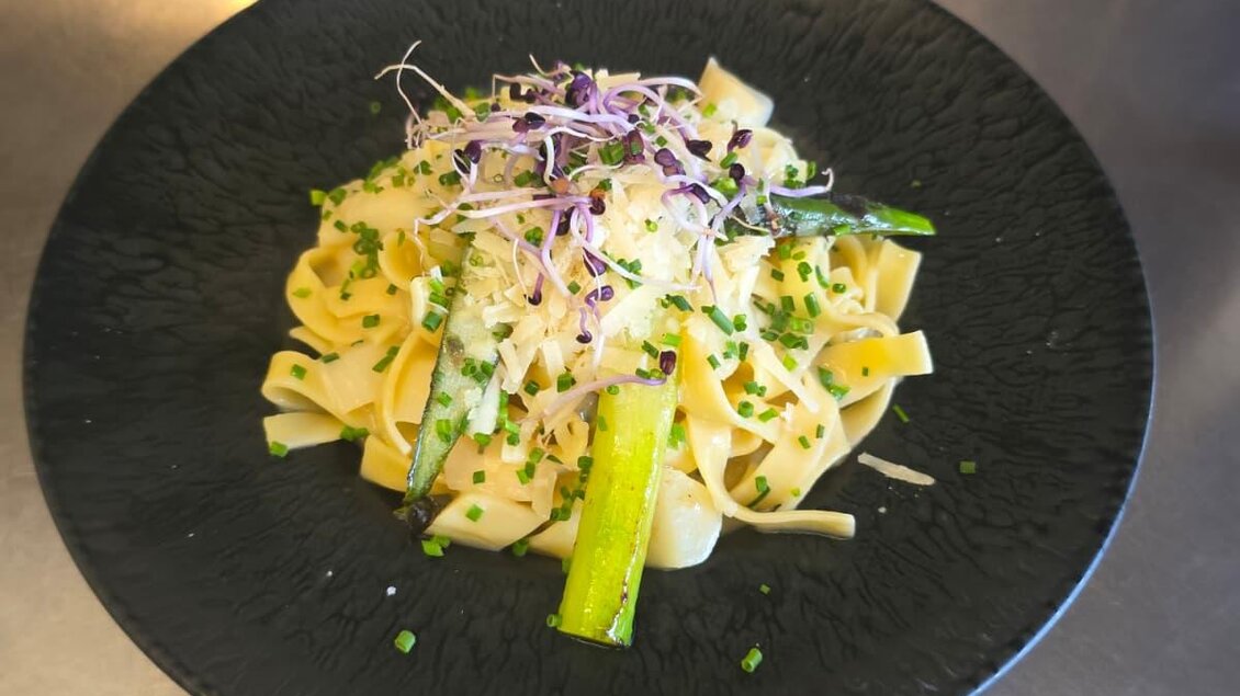 Ein eleganter Teller mit Bandnudeln, garniert mit Spargel und frischen Kräutern. Das Gericht ist ansprechend angerichtet und wirkt frisch und appetitlich.