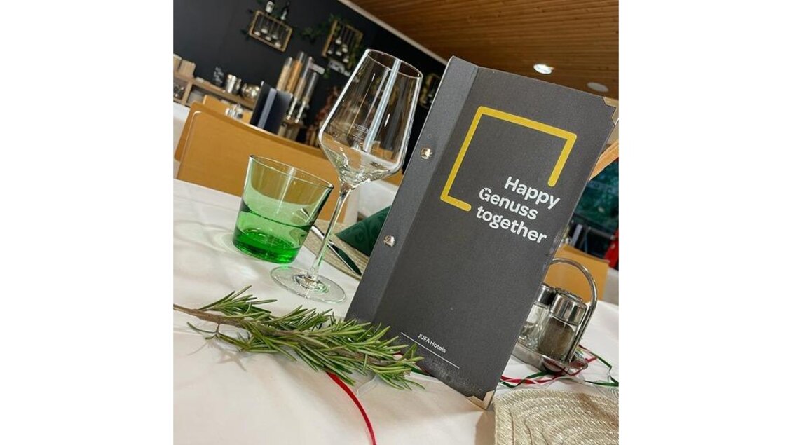 Ein eleganter Tisch in einem Restaurant mit einem Menü, Gläsern und frischen Kräutern. Die Atmosphäre wirkt einladend und gemütlich. | © JUFA Hotels Leibnitz