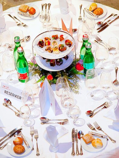Ein schön gedeckter Tisch mit weißen Tischdecken und glänzendem Besteck. Auf dem Tisch stehen Wasserflaschen und ein zweistufiges Dessert-Tablett. | © Revita Gastronomie