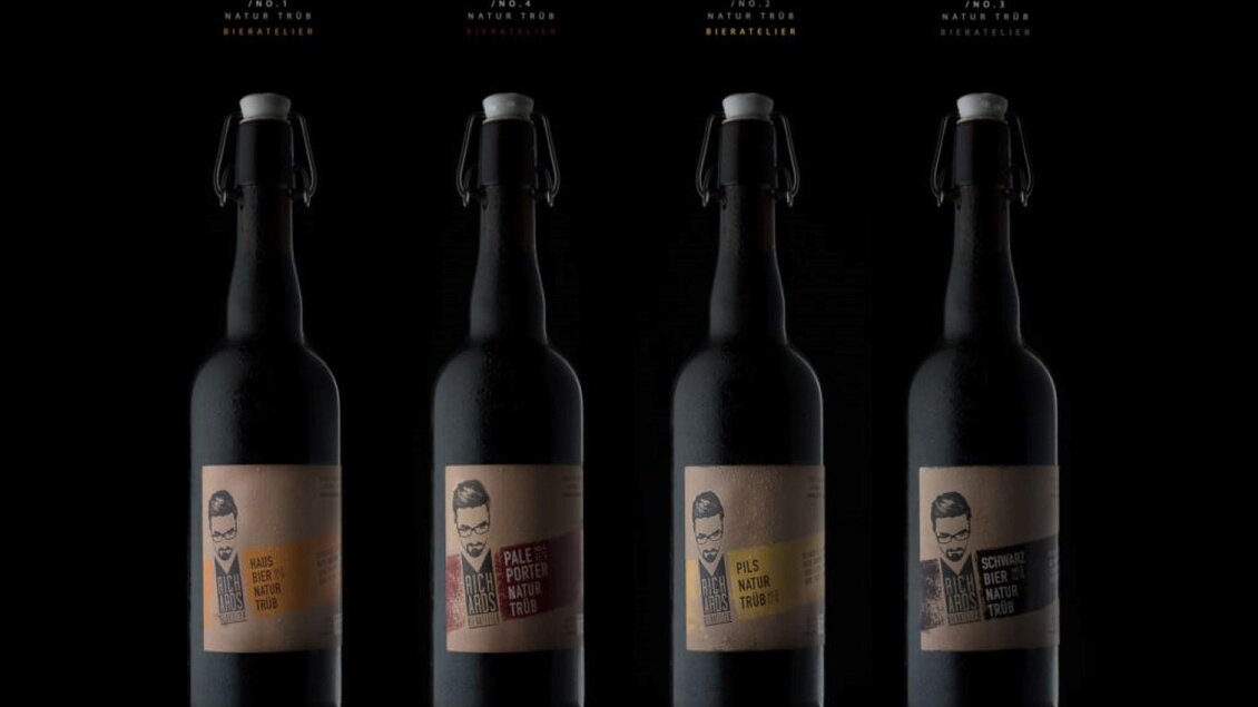 Vier Flaschen mit schwarzem Etikett und verschiedenen Texten stehen vor einem dunklen Hintergrund. Jede Flasche hat ein einzigartiges Design und eine charakteristische Abbildung. | © Richards Bieratelier