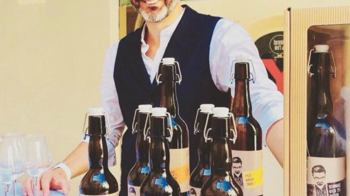 Ein lächelnder Mann steht an einem Tisch mit verschiedenen Flaschen Bier und Gläsern. Im Hintergrund sind einige Verpackungen und Aufkleber zu sehen. | © Richards Bieratelier
