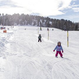 A snow-covered ski area with children skiing. In the background, there are trees and a clear, sunny sky. | © Murtal Seilbahnen Betriebs GmbH