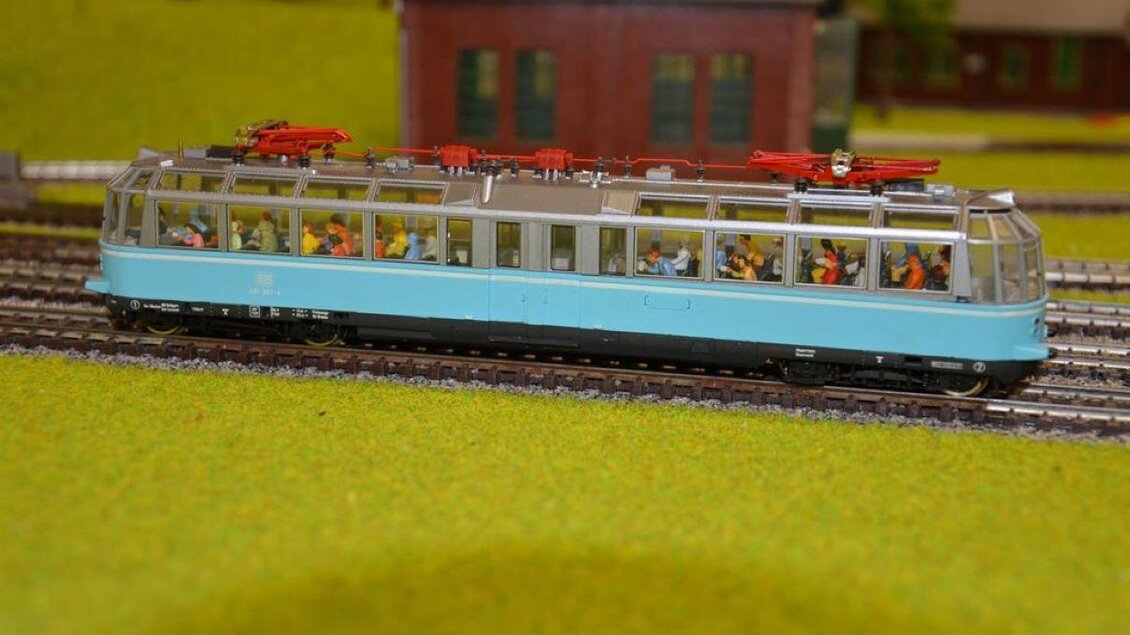 Ein Modellzug in Blau mit vielen Figuren darin fährt auf Schienen. Im Hintergrund sind Gebäude in einer Miniaturlandschaft zu sehen. | © Karl Trummer