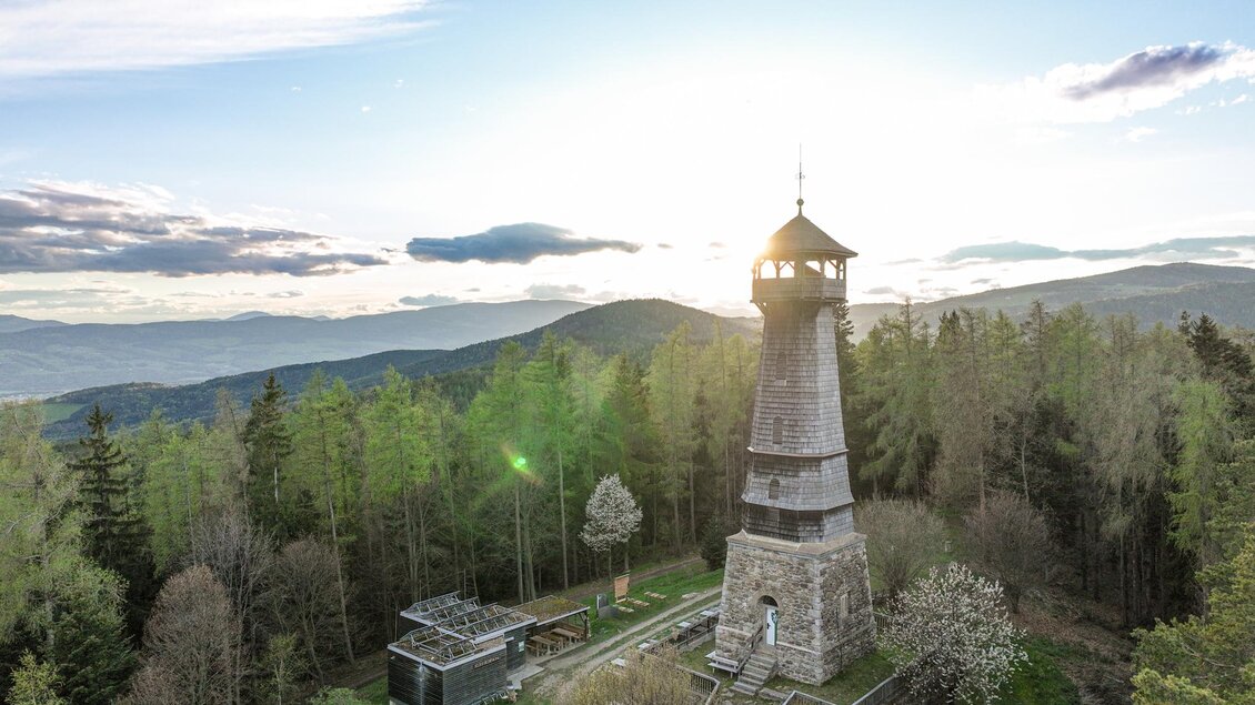 Ein Aussichtsturm inmitten eines Waldes mit sanften Hügeln im Hintergrund. Die Sonne geht hinter den Bergen unter und sorgt für eine malerische Stimmung. | © Oststeiermark Tourismus