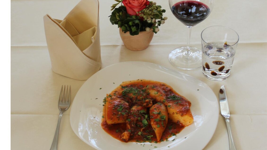 Ein Teller mit Nudeln in Tomatensauce, garniert mit Kräutern. Neben dem Teller steht ein Glas Rotwein und ein kleines Blumengefäß auf dem Tisch. | © Da Gino