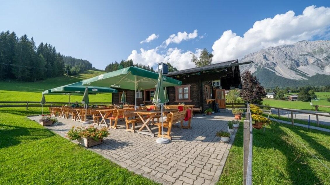 Ein charmantes Bergrestaurant mit Außenbereich und Sonnenschirmen. Im Hintergrund erheben sich grüne Hügel und majestätische Berge unter einem blauen Himmel. | © Alwin Strasser
