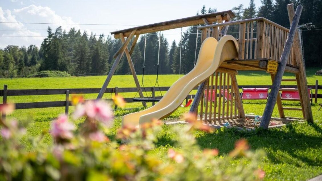Ein Spielplatz mit einer Holzschaukel und einer Rutsche in einer grünen Wiese. Im Hintergrund sind Bäume und ein blauer Himmel zu sehen. | © Alwin Strasser