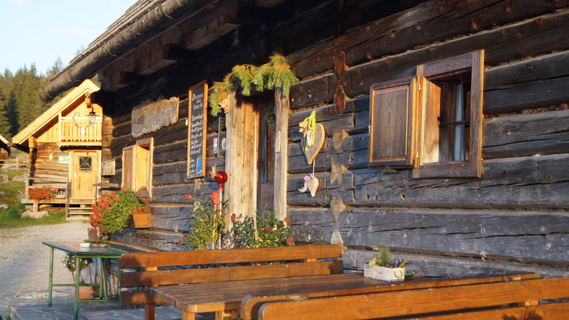 Ein rustikales Holzhaus mit blühenden Pflanzen und offenen Fenstern. Vor dem Haus stehen eine Tischgruppe und Stühle aus Holz. | © Marianne Gruber