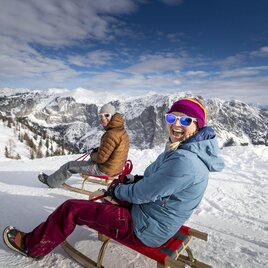 Two people are sitting on sleds in the snow and laughing. In the background, there are snow-covered mountains and a blue sky visible. | © TVB Ausseerland - Salzkammergut-Tom Lamm
