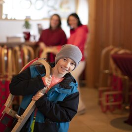 A cheerful boy is carrying a sled and standing in a cozy reception area. In the background, two women can be seen talking. | © TVB Ausseerland - Salzkammergut-Tom Lamm