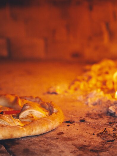 Eine frisch gebackene Pizza liegt in einem Holzofen. Im Hintergrund brennt das Feuer und sorgt für eine gemütliche Atmosphäre. | © Adobe Stock_Pizzeria (1)
