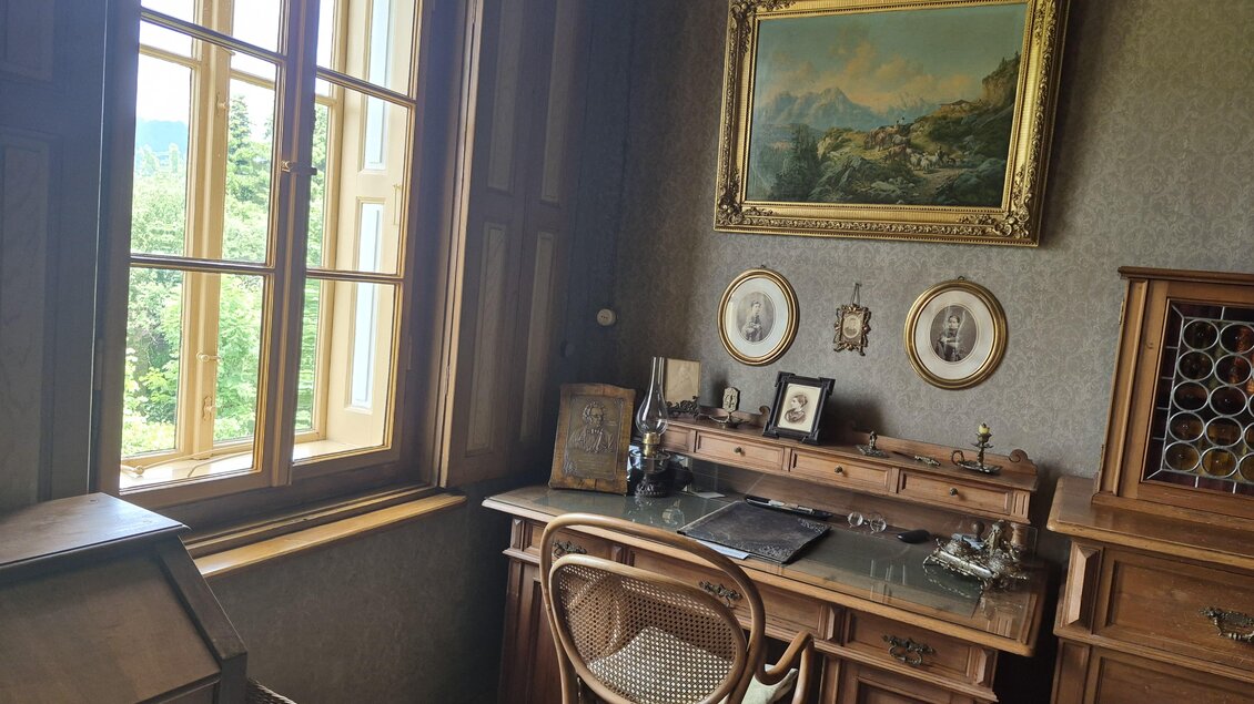 Ein elegantes Arbeitszimmer mit einem Holzschreibtisch und antiken Möbeln. Durch das Fenster fällt viel Tageslicht und bietet einen Blick ins Grüne. | © TV Hochsteiermark/Andrea Stelzer