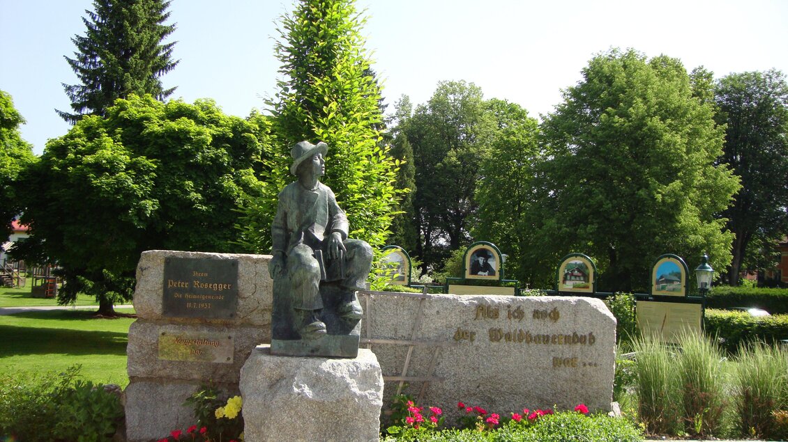 Eine Statue eines Jungen mit einem Hund steht in einem Park. Im Hintergrund sind grüne Bäume und Blumen zu sehen. | © Marktgemeinde Krieglach