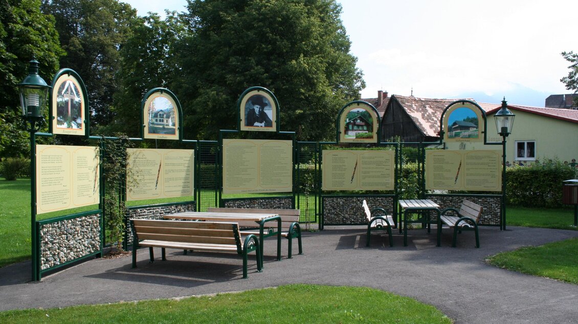 Eine Informationswand mit Tafeln in einem Park. Im Vordergrund steht eine Bank und Tische auf einer Rasenfläche. | © TV Krieglach