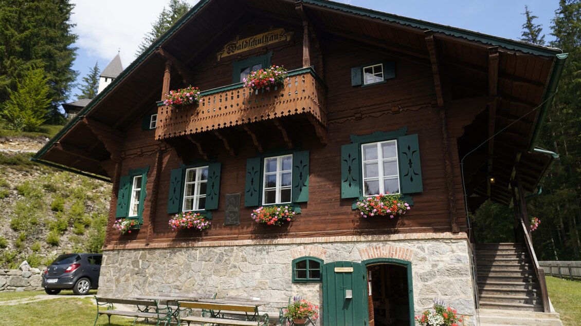 Ein traditionelles Holzhaus mit grünen Fensterläden und Balkonen, geschmückt mit Blumen. Umgeben von Bäumen und einer schönen Landschaft. | © TV Hochsteiermark/Amelie Kertelits