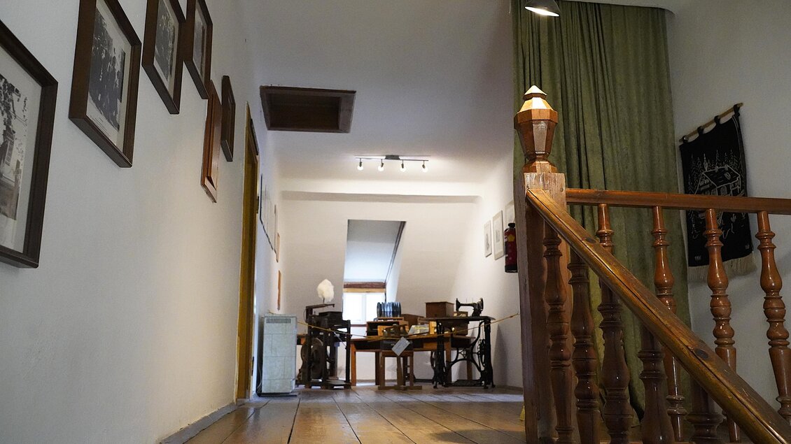 Ein heller Flur mit Holztreppe und Bildern an der Wand. Im Hintergrund sind Tische und Stühle zu sehen. | © TV Hochsteiermark/Amelie Kertelits
