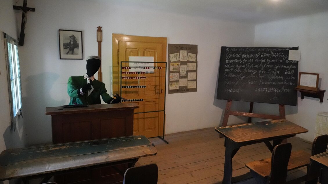 Ein klassischer Schulraum mit Holztischen und einer Tafel. Ein Anzug mit einem Kopf auf einem Pult steht vorne im Raum. | © TV Hochsteiermark/Amelie Kertelits