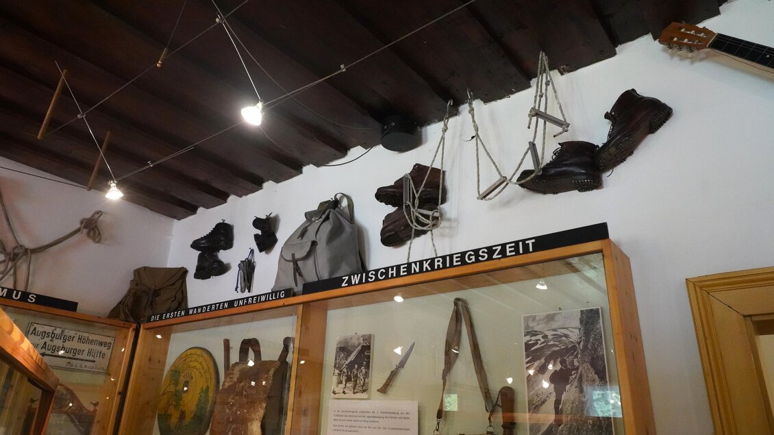 Eine Ausstellung mit historischen Gegenständen aus der Zwischenkriegszeit ist zu sehen. Oben hängen Schuhe, Gurte und andere Ausrüstungsstücke an der Wand. | © TV Hochsteiermark/Amelie Kertelits