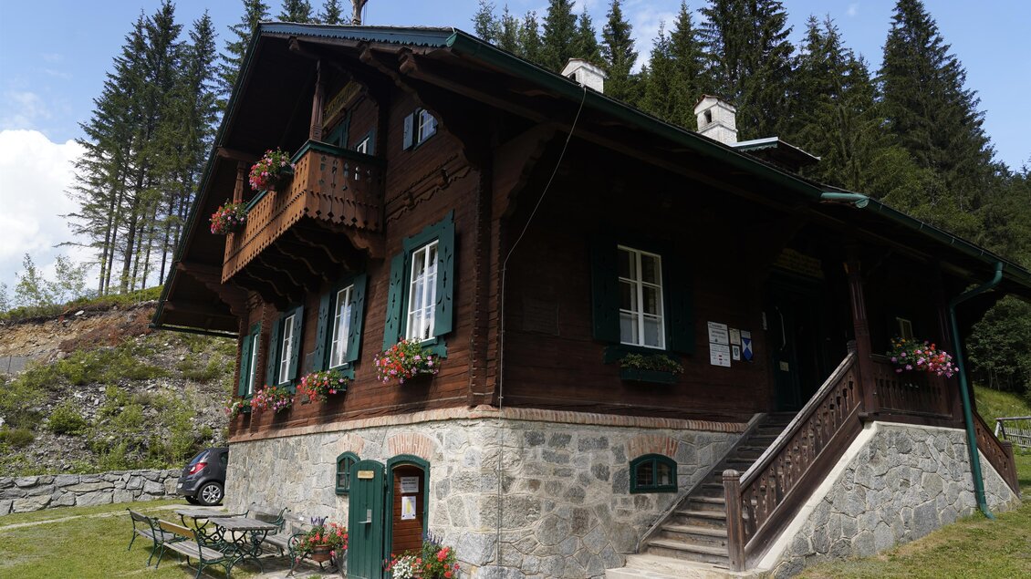 Ein traditionelles Holzhaus mit einem Balkon und Blumenkästen. Es liegt in einer grünen Umgebung mit Bäumen im Hintergrund. | © TV Hochsteiermark/Amelie Kertelits