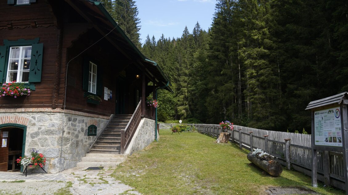 Ein charmantes Holzhaus neben einem Weg, umgeben von dichten, grünen Wäldern. Der Himmel ist klar und die Atmosphäre ist ruhig und einladend. | © TV Hochsteiermark/Amelie Kertelits